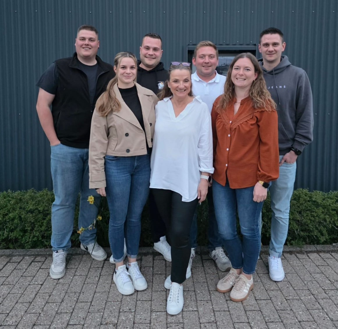 Sieben Personen stehen gemeinsam draußen vor einer dunklen Wand und schauen in die Kamera. Sie sind locker nebeneinander und leicht versetzt angeordnet, tragen Alltagskleidung und wirken freundlich.