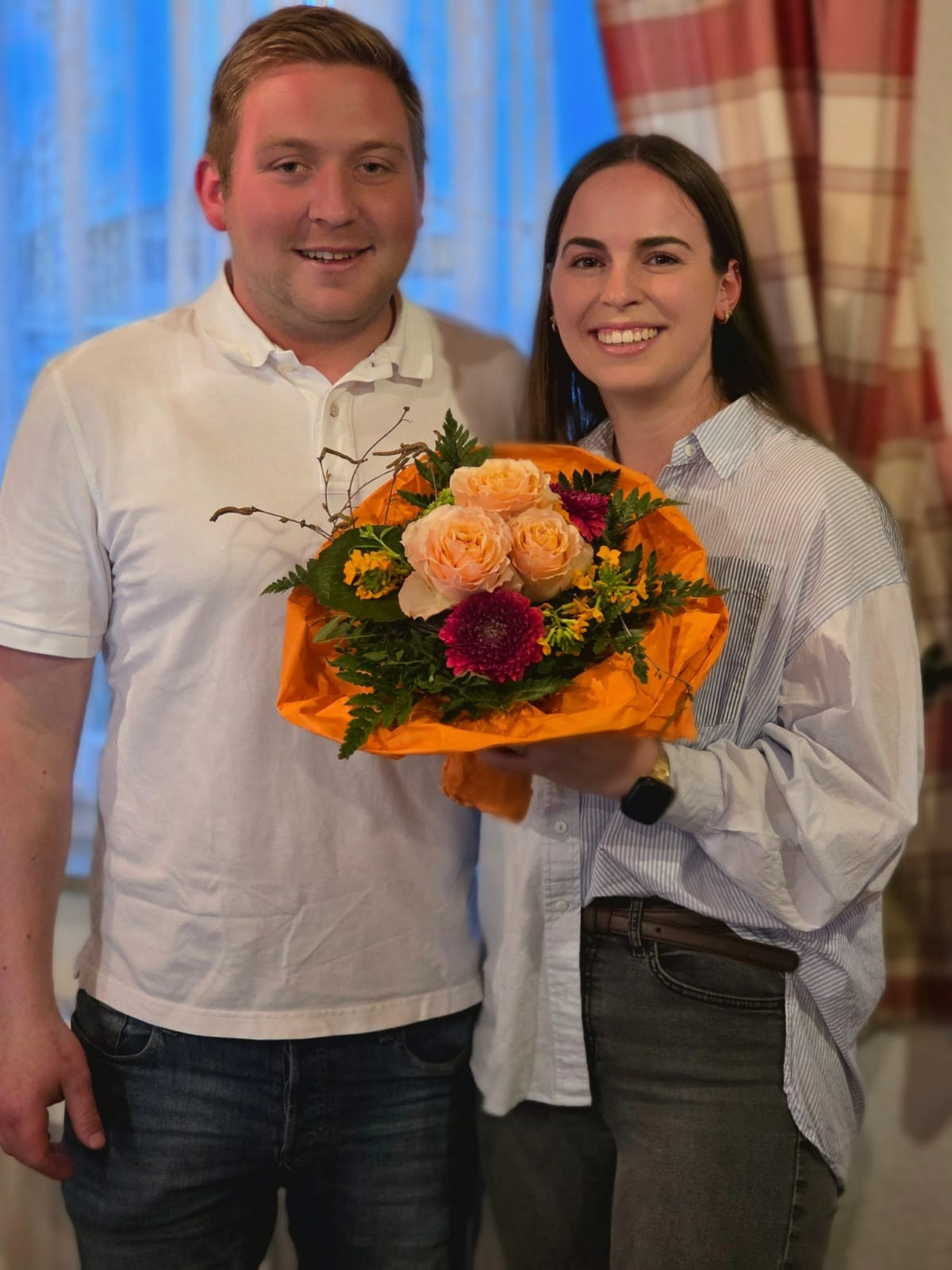 Sieben Personen stehen gemeinsam draußen vor einer dunklen Wand und schauen in die Kamera. Sie sind locker nebeneinander und leicht versetzt angeordnet, tragen Alltagskleidung und wirken freundlich.